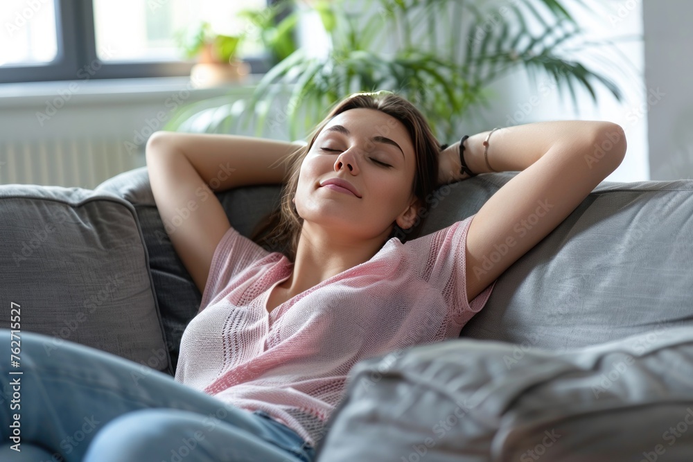 A woman is laying on a couch, looking relaxed and peaceful. The couch is covered in a gray fabric, and there is a potted plant in the background A woman is laying on a couch, looking relaxed and peaceful. The couch is covered in a gray fabric, and there is a potted plant in the background
