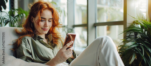 
A smiling professional woman with red hair and green eyes lounges on an office sofa, holding a yellow phone, gazing through a window at a plant-filled modern interior.