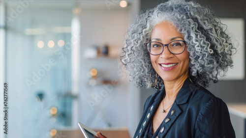 
A professional woman in her mid-40s with curly gray hair and cinematic makeup smiles, holding an iPad near her work desk with a laptop, glasses on, in business attire, against a backdrop of modern gl photo