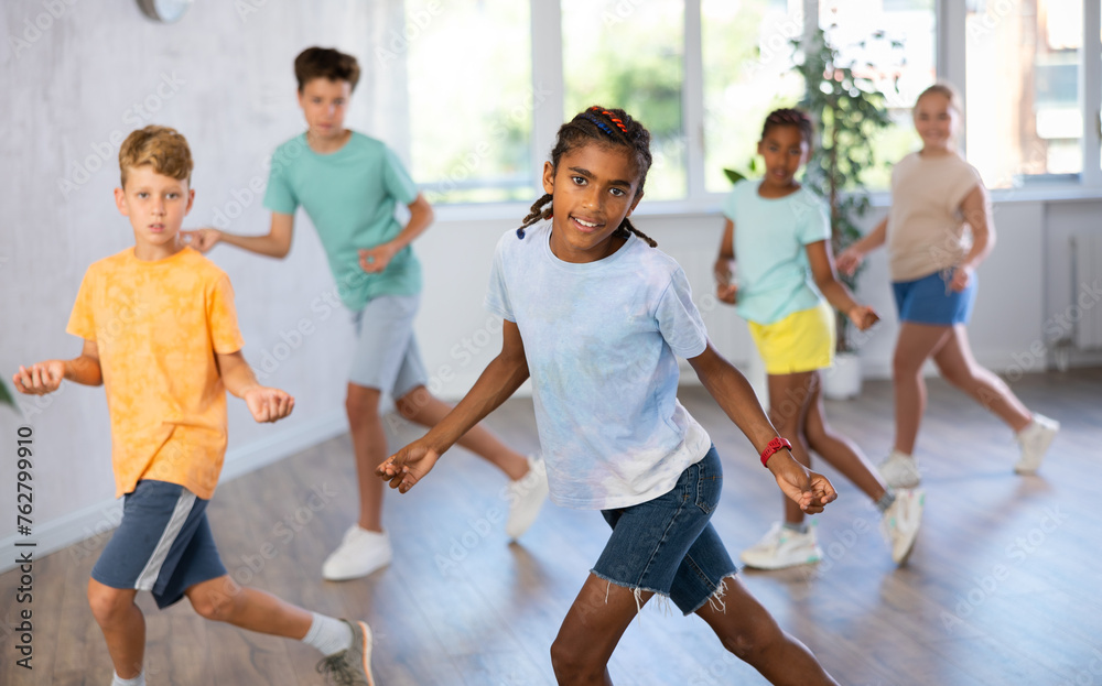 Fototapeta premium African boy performs movements during warm-up, limbering-up part of workout together with peers. Group of young girls and guys dance modern waacking in fitness club unfocused