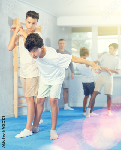 Concentrated teen boys practicing armlock, painful control move to hold and immobilize opponent, in training bout during self defence course with male trainer in gym ..