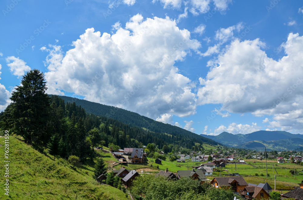 Serene village landscape with houses nestled on the valley among rolling green hills with mountain ridge on background under blue sky with clouds. Verkhovyna, Carpathian Mountains, Ukraine