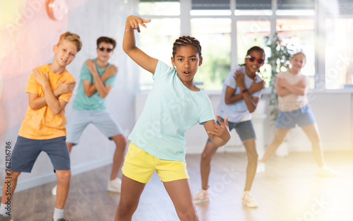 African female child perform choreographic exercises and teaches energetic mobile social dance waacking together with friends. Young girls and guys repeat movements, train in spacious studio unfocused