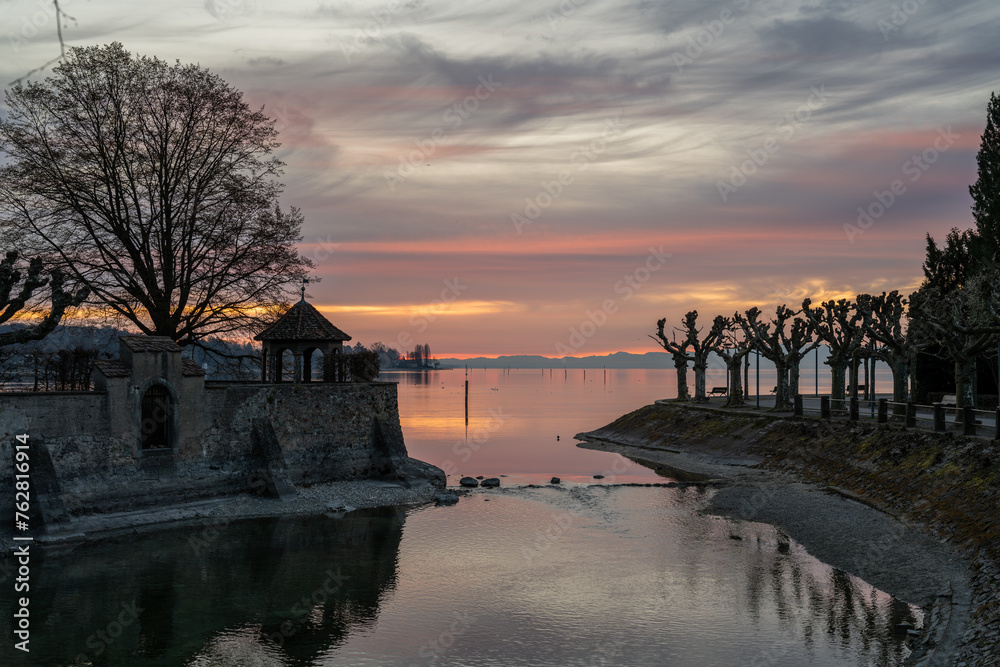 Morgendämmerung über dem See beim Wasserkanal an der Steinmauer am Steigenberger Hotel mit den Bergen am Horizont. Konstanz, Bodensee, Baden-Württemberg, Deutschland, Europa.