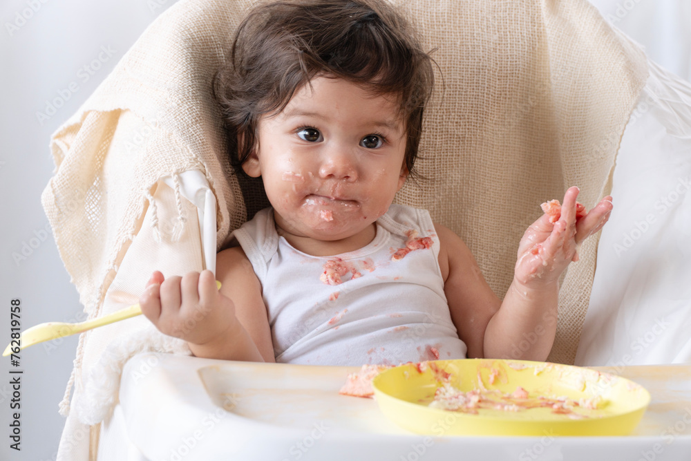 Innocence toddler kid sloppy mouth hand eating birthday cake sitting on chair at home. Happy mixed race children daughter with sweet birthday cake messy face enjoyment sitting chair. Growing life. Innocence toddler kid sloppy mouth hand eating birthday cake sitting on chair at home. Happy mixed race children daughter with sweet birthday cake messy face enjoyment sitting chair. Growing life.