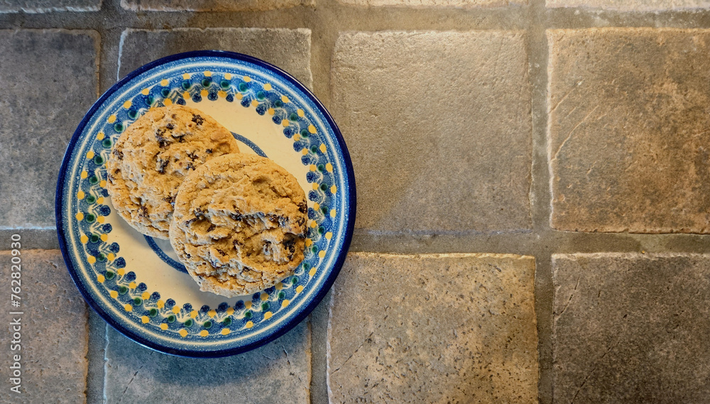 two oatmeal raisin cookies on a floral design plate on a porcelain tiled counter top