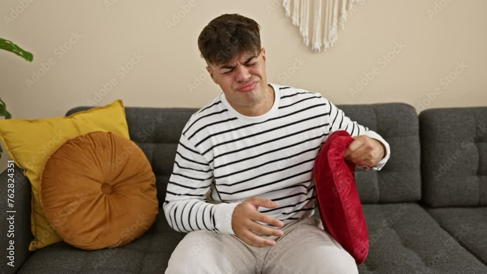 Worried young hispanic man suffers stomachache, unhappily lying on home sofa, hugging cushion, in room's indoor interior, indicating serious sickness