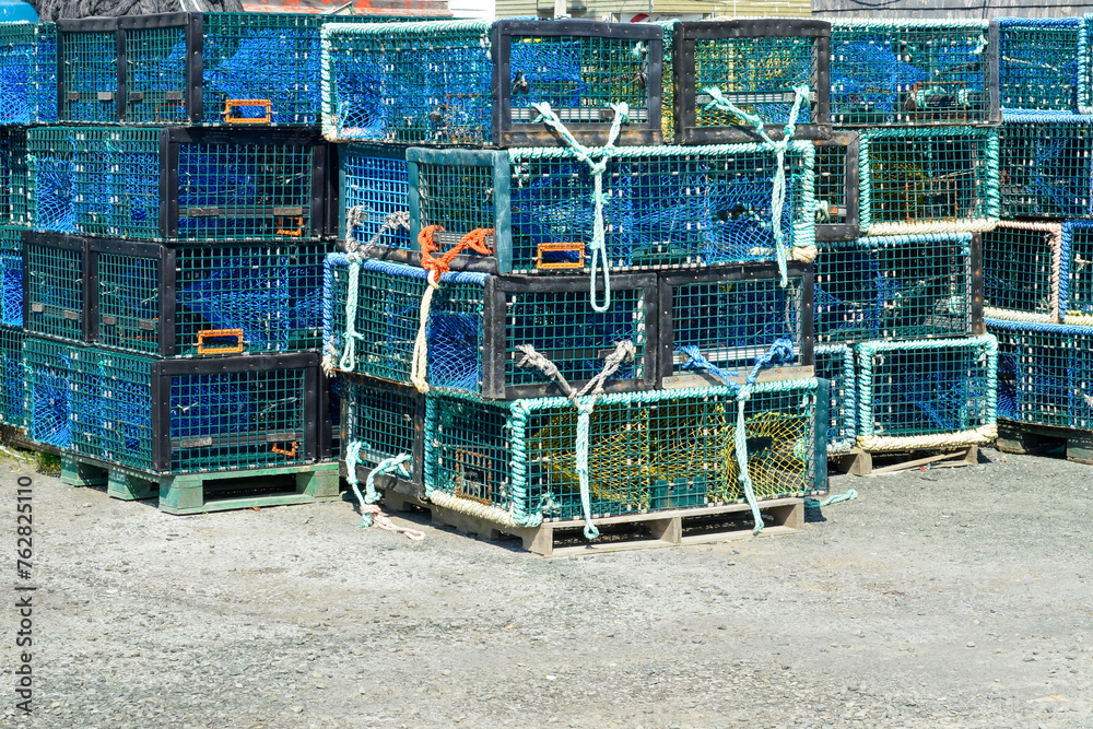 Multiple yellow lobster pots or traps stacked high along a wharf. Containers are made from yellow wire and nylon netting and in a square shape. There's no bait in the pots, all are empty.