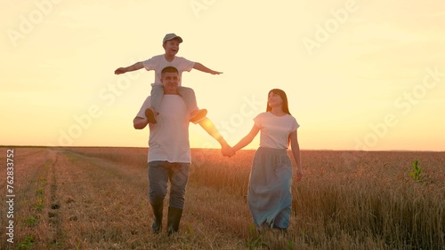 Happy farmers family with child go through wheat field sun. Full family, people in nature, holiday. Child son on shoulders of dad, mom, walk hand in hand outdoor. Parental care for child. Dream of fly