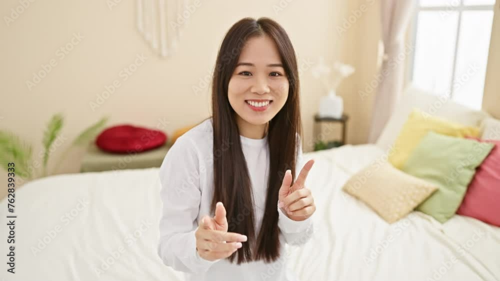 A cheerful young asian woman in a white shirt poses indoors in a bright, cozy bedroom setting with colorful pillows.