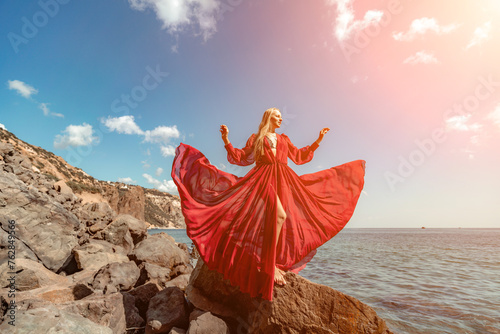 Red dress sea woman. A blonde with flowing hair in a long red dress sits on a rock near the sea. The concept of trips, a photo shoot at the sea