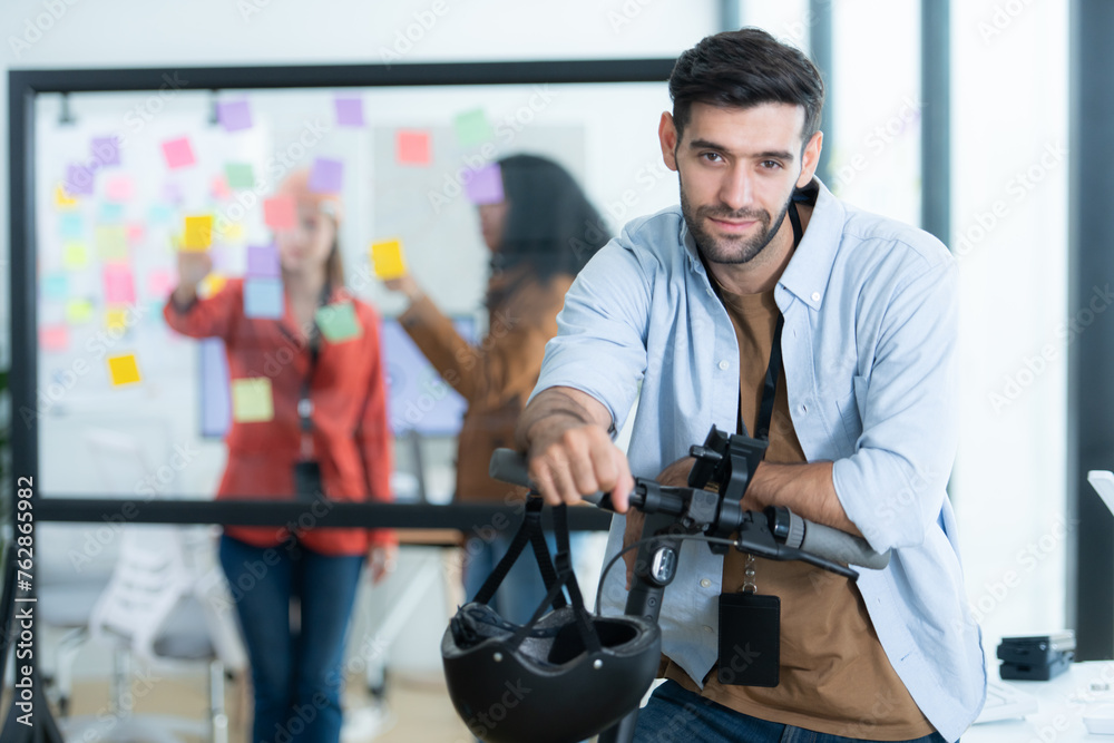 Portrait of smiling businessman riding electric scooter in office with colleague in background