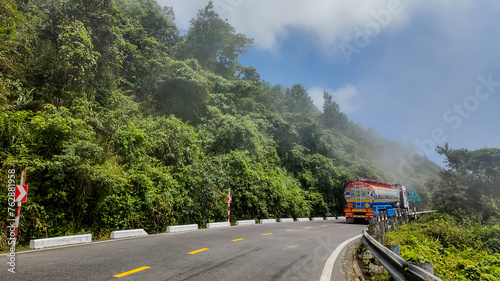 Colorful truck driving on a winding mountain road surrounded by lush greenery with fog in the background, suitable for travel and transportation themes with copy space