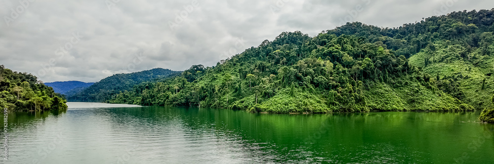 Panoramic view of a tranquil tropical lake with lush green rainforest mountains under a cloudy sky, ideal background for eco-tourism and nature conservation themes, Earth day concept