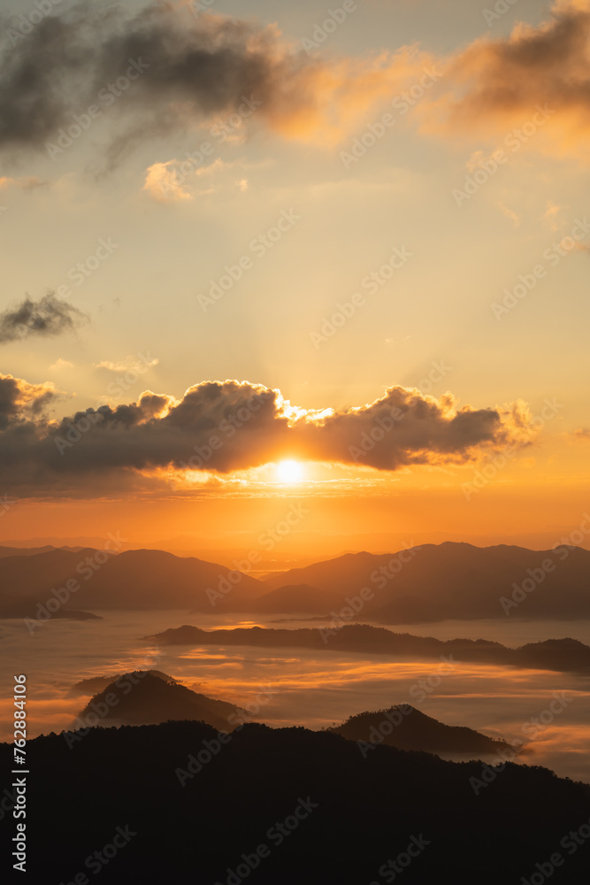 The sun is setting over a mountain range, casting a warm glow over the landscape. The sky is filled with clouds, creating a sense of depth and atmosphere. The mountains are covered in a mist