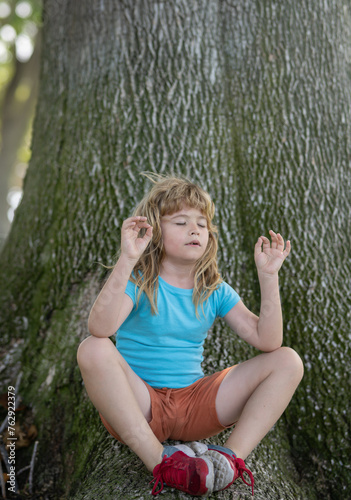 Peaceful cute little boy holding fingers in mudra gesture and meditating with closed eyes, feeling calm positive and relaxed on nature background. Kids yoga practice.