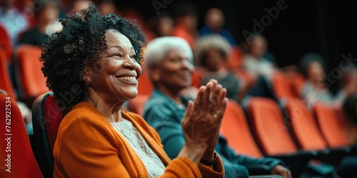 Senior African American woman enjoying a live performance in a theater, sitting comfortably in a plush seat and expressing joy through applause with a radiant smile on her face