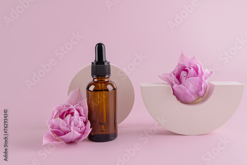 Glass bottle with natural cosmetics, serum, oil for face and body skin care on the background of concrete geometric shapes and flowers.