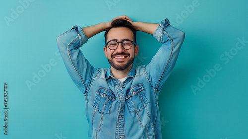 Portrait of satisfied good-looking european male in glasses and denim shirt, holding hands behind head, smiling broadly and gazing at camera with pleased expression, standing on teal color background photo