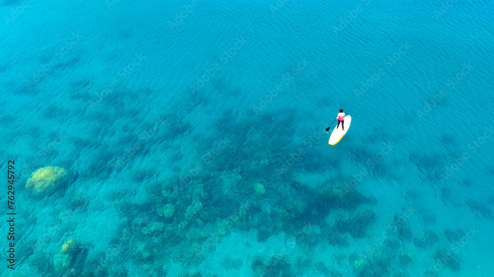 Aerial view of a woman paddle boarding in the blue sea. Clear water. You can see the coral beneath the sea.
