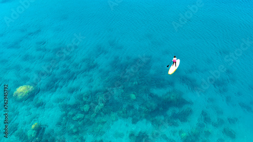 Aerial view of a woman paddle boarding in the blue sea. Clear water. You can see the coral beneath the sea. © Photo Sesaon