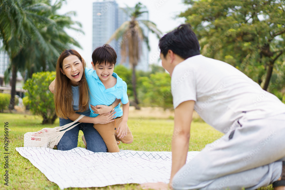 Happy cheerful Asian family with father, mother, and little son enjoy picnic together in a weekend at a park.