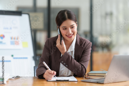 Asian businesswoman is talking on her cell phone while writing on a clipboard © amnaj