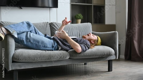 A guy is lounging on the couch at home and throwing corn sticks into the air to catch them in his mouth.