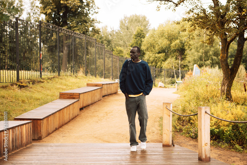 Portrait of a young african man dressed in a blue sweatshirt, holding a backpack, walking in the park on a summer day
