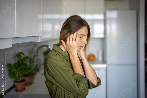 Tired woman feeling headache rubbing temples standing on kitchen with closed eyes at home. Blonde female suffering from migraine head pain. Having headache cephalalgia premenstrual syndrome feel bad. 