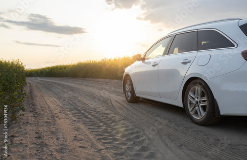 white car drives along a country road among a blooming rapeseed field towards the setting sun towards adventure. Travelling by car. Weekend trip