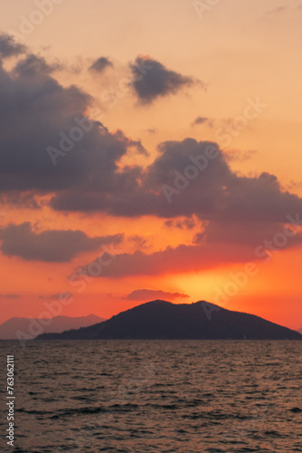 As the sun sets over the ocean, the sky transforms into a canvas of orange and red hues, casting a warm afterglow over the water and the distant mountain