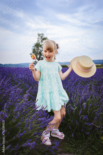 smiling little girl holding straw hat and lollipop behind her lavender field