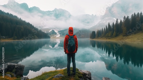 Traveler with red hat standing before a serene mountain lake with misty peaks in the background.