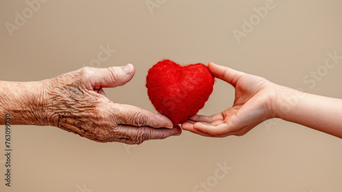 A plush red heart is exchanged between a wrinkled elderly hand and a smooth young hand.