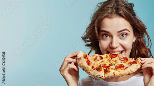Attractive cheerful woman eats delicious Italian pizza  surprised with wonderful taste  on pastel blue background