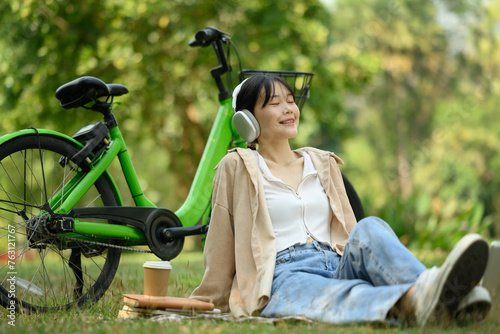 Happy young woman listening to music with headphone sitting at the park with her bicycle