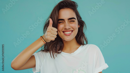 Beautiful brunette woman wearing casual clothes winking and showing thumbs up at camera on pastel blue background