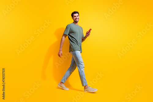 Full length photo of cheerful good mood man dressed khaki t-shirt texting messages modern gadget isolated yellow color background
