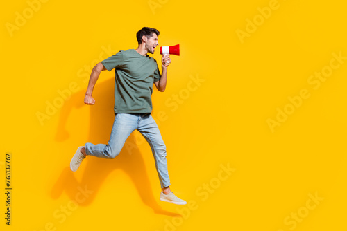 Full length photo of excited cheerful guy wear khaki t-shirt jumping high screaming bullhorn empty space isolated yellow color background