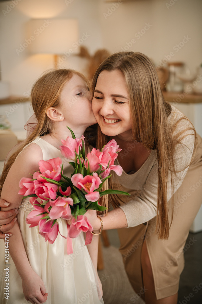Daughter kisses and hugs mom. Happy family holiday. Cute little child girl greets smiling mother in kitchen. Beautiful kid gives mother big bouquet of tulip flowers and makes surprise for Mother's Day Daughter kisses and hugs mom. Happy family holiday. Cute little child girl greets smiling mother in kitchen. Beautiful kid gives mother big bouquet of tulip flowers and makes surprise for Mother's Day