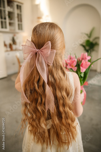 Beautiful girl with long hair and big bow on her head is standing with her back and holding a big bouquet of tulips. The daughter gives mother flowers and makes surprise for Mothers Day. Back view