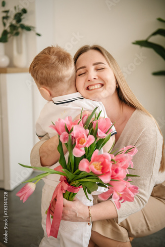 Son hugs smiles mom. Child makes surprise for Mothers Day. Back view. Little boy holds, gives bouquet of tulip flowers, congratulates mommy in kitchen. International Women's Day. Happy family holiday.