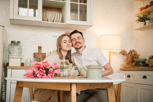 Loving couple having conversation and drinking coffee in morning in kitchen. Romantic male hug female, spending time together. Man congratulate woman and give bouquet of flowers on Women's day.