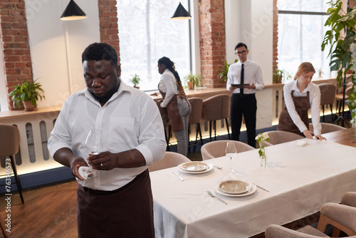 High angle view selective focus shot of young Black waiter and his multi-ethnic colleagues cleaning glassware and setting tables in modern restaurant