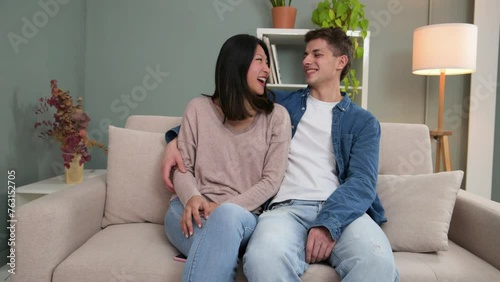 Diverse carefree man and woman hugging and relaxing together in couch at the living room. Affectionate smiling young adult couple sitting on sofa at home looking to camera.