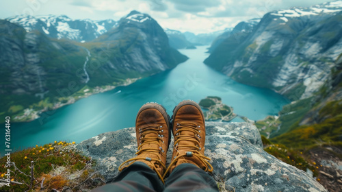 A traveller sits at the edge of a high rocky ledge, legs extended, showing only their brown hiking boots with the stunning backdrop of a nature