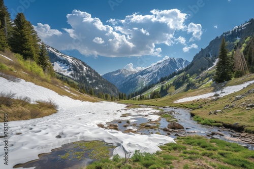 Spring thaw in a mountain valley featuring a partially frozen stream, melting snow, green grass patches, and towering spruce trees under a sunny sky with cumulus clouds