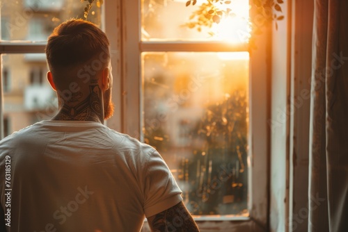 Man in a fitted t-shirt, silently observing the sunset through a window. His arms, decorated with elegant tattoos, are highlighted by the golden hour light, emphasizing the beauty of the moment.