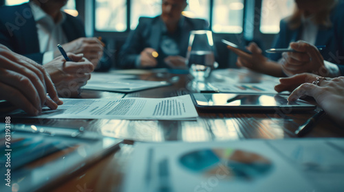 Close-up of a business meeting in progress, focusing on documents with charts and pens on a table with partial view of participants engaged in discussion in a dimly lit room.